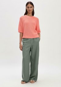 Maglione a maniche corte in corallo con un motivo texturizzato, abbinato a pantaloni larghi verde oliva e sandali beige chiaro.