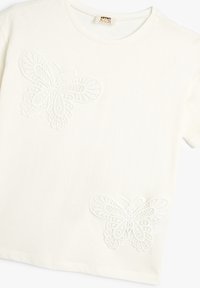 Witte katoenen t-shirt met twee geborduurde vlinders in een kantontwerp, gepositioneerd aan de voorkant, met korte mouwen en een ronde hals.