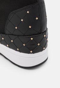 Czarne sneakersy z teksturowanej siateczki i pikowanymi skórzanymi akcentami, z złotymi ćwiekami na pięcie i kontrastującą białą gumową podeszwą.