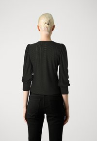 Top negro texturizado con mangas abullonadas, que presenta un patrón geométrico. Combinado con unos jeans negros, que muestran bolsillos y una silueta ajustada.