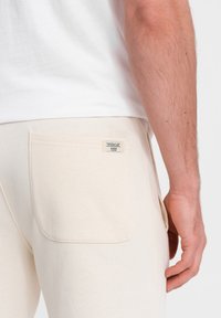 Beige Baumwoll-Jogginghosen mit einer aufgesetzten Tasche und einer Label-Angabe. Sichtbare Nähte und weicher Stoff. Weißes T-Shirt, das an der Taille übereinanderliegt.