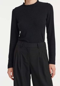 Schwarzes, langärmeliges, eng anliegendes Top mit Rüschensaum, kombiniert mit hoch sitzenden, gefalteten, weiten schwarzen Hosen an einer Person.