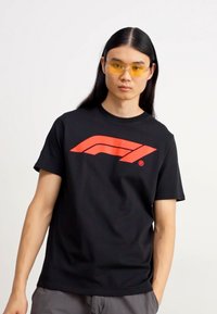 Tricou negru cu un logo grafic roșu îndrăzneț. Mâneci scurte, design cu guler rotund, confecționat dintr-un material moale. Modelul poartă ochelari de soare galbeni.