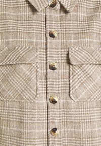 Camisa de cuadros beige, mezcla de lana, con grandes bolsillos en el pecho, cierre de botones y cuello redondeado. Tejido texturizado con rayas blancas en contraste.