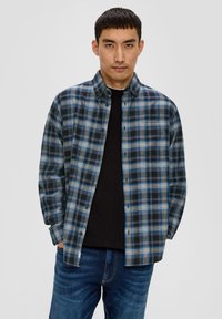 Chemise à boutons en bleu, noir et blanc à carreaux. Présente un col classique et des manches longues, portée sur un pull texturé noir.