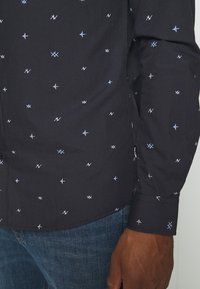 Camicia button-up blu scuro con motivi geometrici bianchi e azzurri. Dettagli notevoli includono un orlo curvo e polsini con un solo bottone.