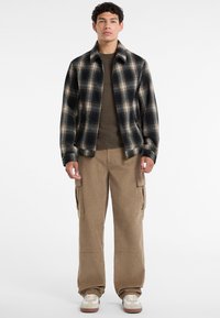 Karirano flanel kožuh u tamnoplavoj i bež boji, dizajn s kragnom, u kombinaciji s bež cargo hlačama koje imaju bočne džepove i opušteni kroj.