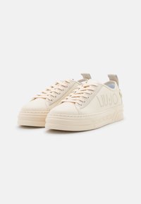 Un paio di sneaker basse beige con suole spesse, lacci e dettaglio del logo sul lato, posizionate su uno sfondo bianco.