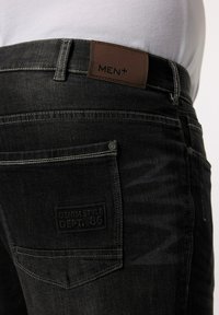 Svarta denimjeans med en läderlapp märkt "MEN+". Funktioner inkluderar synlig sömnad, en bakficka med varumärkeslogotyp och en figursydd design.