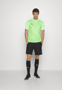 Zaļš rakstains īso piedurkņu krekls, melni šorti, melni līdz ceļiem augsti zābaki, pabeigti ar melniem futbola apaviem un Puma logotipa akcentiem.