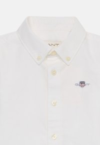 White shirt with a button-down collar, pearlescent buttons, and an embroidered GANT logo on the chest. Smooth cotton fabric texture.