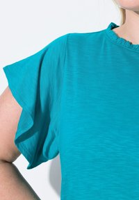Tyrkysový top s krátkým rukávem, ozdobený jemným volánkovým výstřihem a měkkou, hladkou texturou; má volný střih a lehký materiál.