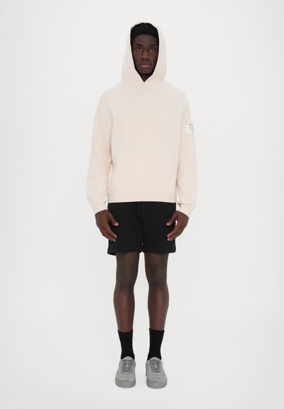 Hellbeige Kapuzenpullover mit Kängurutasche, kombiniert mit schwarzen Shorts. Graue Sneakers und schwarze Socken runden das Outfit ab.