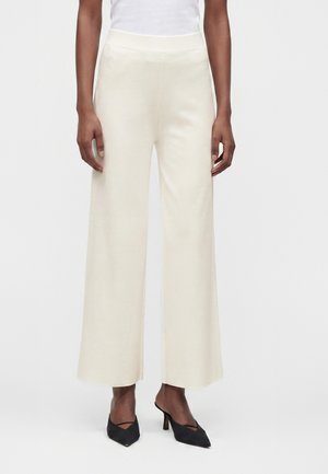 Femme portant un pantalon crème taille haute à jambes larges, associé à des chaussures à talons noires à bouts pointus, debout devant un fond uni.
