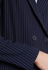 Dvojřadý námořnický blazer s bílými proužky, se dvěma černými knoflíky a strukturovanou tkaninou. Detail rukávu a límce zblízka.