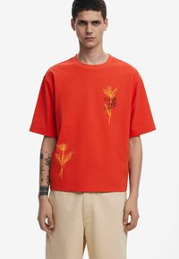 Rød kortærmet t-shirt lavet af bomuld, med gule broderede blomsterdesigns og tekst, i en afslappet pasform og rund halsudskæring.