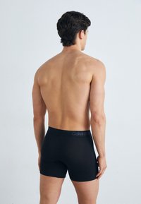 Boxers noirs avec une ceinture élastique douce arborant un logo violet. Modèle montré de dos, avec un design lisse et épuré.