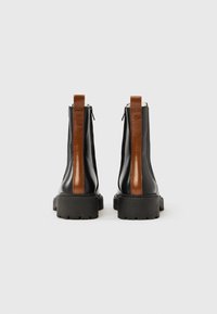 Botas de cano curto em couro preto com uma faixa brilhante castanha, sola grossa de borracha e fechos laterais, exibindo um design elegante e moderno.