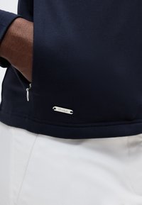 Χέρι στην τσέπη ενός ναυτικού μπλε μπουφάν με ασημένιο φερμουάρ και μικρή μεταλλική ετικέτα μάρκας, συνδυασμένο με λευκό παντελόνι.