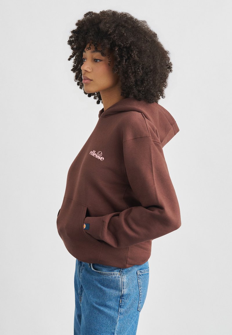 Femme aux cheveux bouclés portant un sweat à capuche marron de la marque ellesse et un jean bleu, debout de profil avec les mains dans la poche du sweat, sur un fond uni.