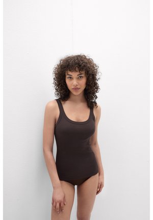 Frau mit lockigem Haar, die ein dunkelbraunes Tanktop und passende Unterwäsche trägt, steht vor einer weißen Wand und schaut nach vorne.