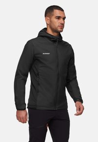 Homme portant une veste à capuche Mammut noire et un pantalon noir, se tenant avec les bras détendus devant un fond blanc uni.