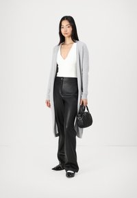Grauer langer Cardigan, weißes tief ausgeschnittenes Top, schwarze Lederhose, schwarze Handtasche, flache Schuhe mit Verzierungen, minimalistisches Design, glatte Texturen.