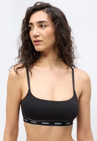 Γυναίκα με σγουρά σκούρα μαλλιά που φοράει ένα μαύρο bralette με το λογότυπο Hugo, κοιτάζει προς τα αριστερά της, πάνω σε ένα απλό φωτεινό φόντο.