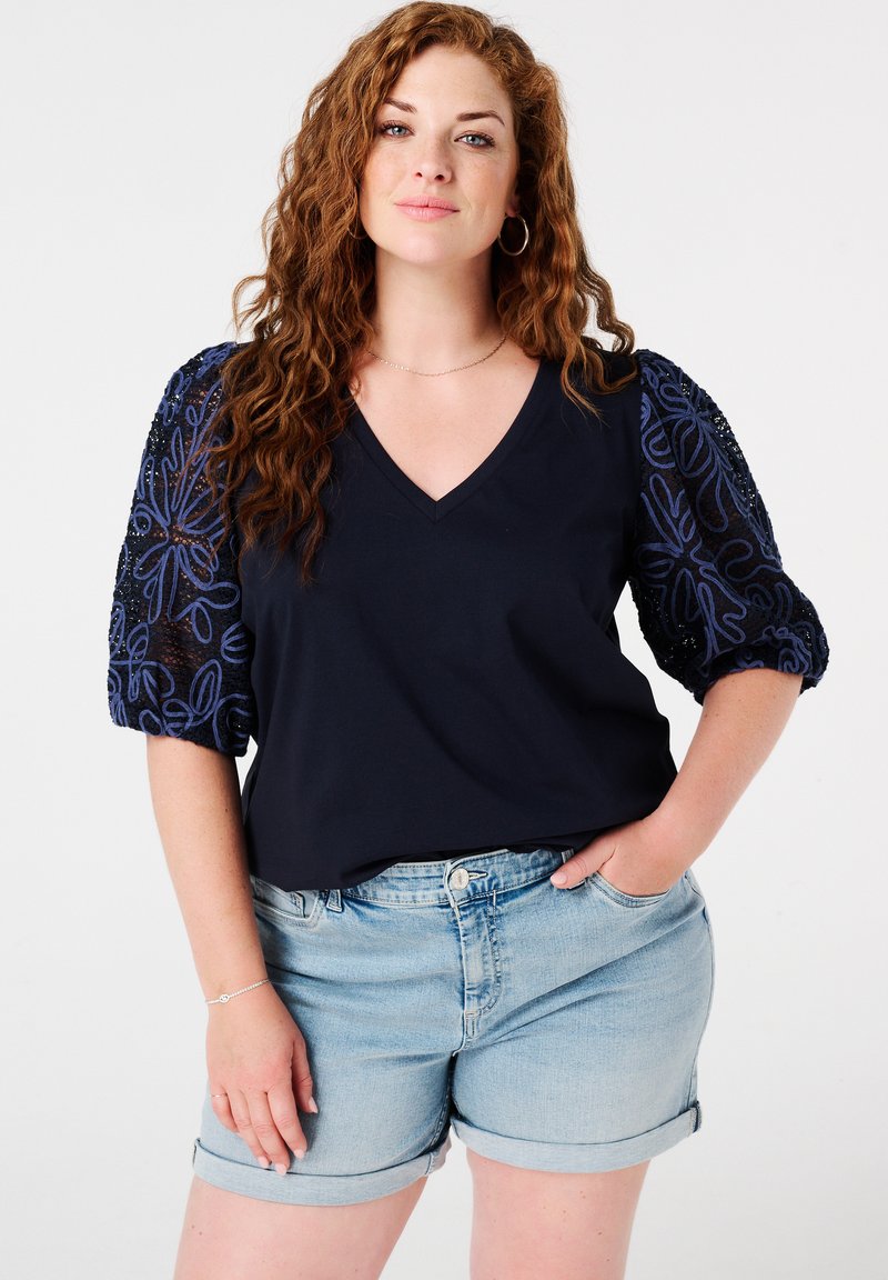 Marineblauw V-hals top met textuur en blauw patroon op de pofmouwen, gecombineerd met lichte denim shorts. Heeft een ontspannen pasvorm en zacht materiaal.