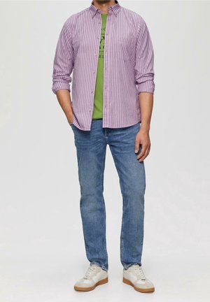 Hombre con camiseta verde estampada, camisa morada de rayas desabrochada, vaqueros azules y zapatillas blancas, de pie con una mano en el bolsillo.