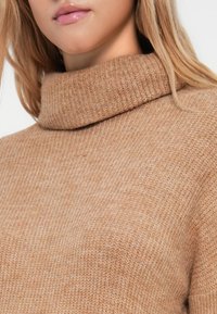 Beige coltrui met een textuur gebreid patroon, stevige kraag en zachte stof. Close-up benadrukt de warme kleur en details.