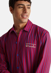 Mladý muž se usmívá, má na sobě červenou a modrou pruhovanou košili s knoflíky a logem "Stranger Colors of Benetton" na hrudi.