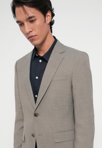 Ανοιχτό γκρι σακάκι με υφή, κλείσιμο με ένα καφέ κουμπί και καμπυλωτές πέτες, φορεμένο πάνω από ένα σκούρο πουκάμισο με κουμπιά.