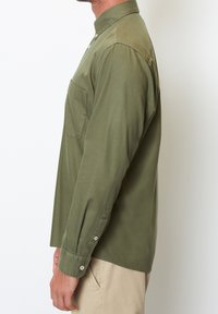 Olivově zelená košile s dlouhým rukávem z měkké látky, s límcem na knoflíky, jednou náprsní kapsou a zapínacími manžetami, nošena s světle béžovými kalhotami.