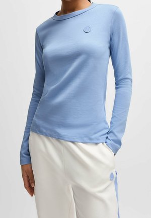 Femme portant une chemise à manches longues bleu clair et un pantalon blanc avec des bandes latérales bleues et des badges circulaires sur la chemise et le pantalon.