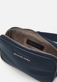 Niebieska tkaninowa torebka na ramię ma zamek błyskawiczny, złote okucia i wnętrze z oznaczeniem "Michael Kors". Gładka tekstura i prostokątny kształt.