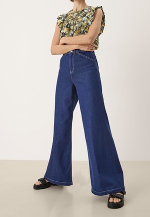 Person iført blå jeans med høj talje og vide ben, blomstret ærmeløs bluse med flæser og sorte platformsandaler stående med armene krydset.