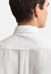 Plecy i szyja mężczyzny z krótkimi ciemnymi włosami, ubrany w białą koszulę z kołnierzykiem i guzikiem na kołnierzyku.