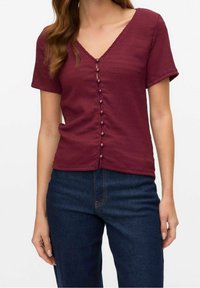 Burgunderfarbene Kurzarmbluse mit V-Ausschnitt, strukturiertem Stoff und einer Reihe kleiner Knöpfe an der Vorderseite. Kombiniert mit dunklen Jeans.