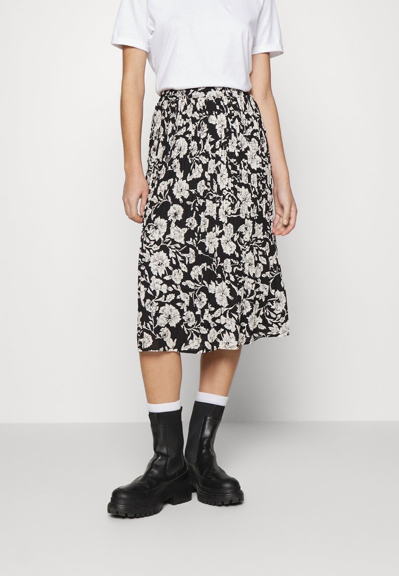 Personne portant une jupe midi fleurie noire et blanche, une chemise blanche à manches courtes, des chaussettes blanches et des bottines noires épaisses, debout sur un sol clair.
