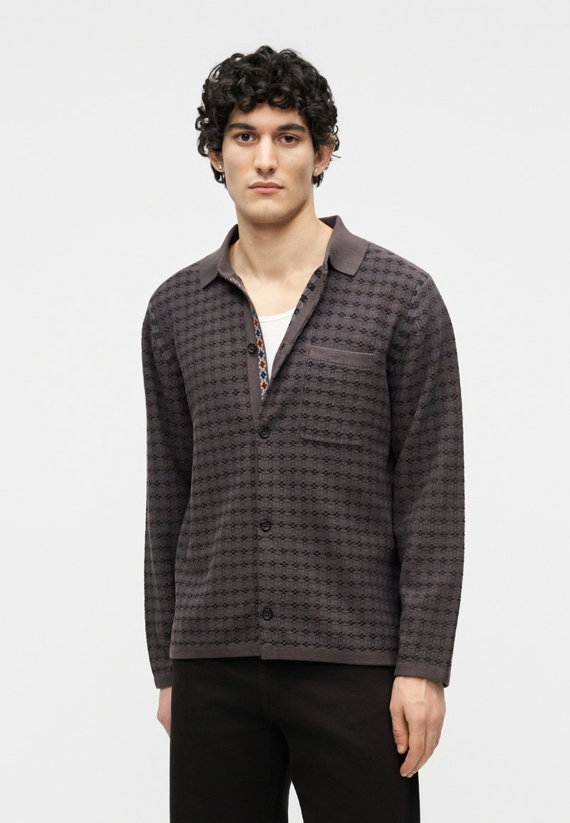 Νεαρός με σγουρά μαλλιά φοράει πουκάμισο με κουμπιά σε σκούρο γκρι με σχέδιο πάνω από λευκό μπλουζάκι και μαύρο παντελόνι, στέκεται μπροστά σε λευκό φόντο.