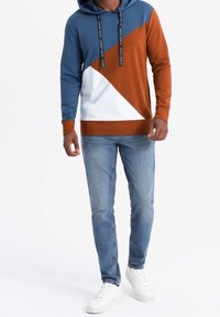 Sudadera con bloque de color en azul, marrón y blanco; mezcla de algodón, con un bolsillo canguro y capucha con cordón. Combinada con jeans de color azul claro y zapatillas blancas.
