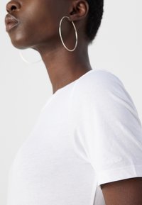 Profil d'une personne portant une chemise blanche unie à manches courtes et de grandes boucles d'oreilles créoles argentées sur un fond clair uni.