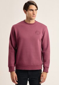 Sudadera burdeos con cuello redondo y puños acanalados. Presenta un parche de logo tonal en el pecho y un dobladillo recto. Fabricada en una suave mezcla de algodón.