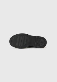 Semelle de chaussure en caoutchouc noir avec un motif texturé, présentant des rainures horizontales et un logo circulaire près du talon.