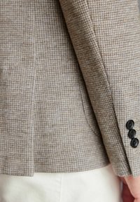 Brauner Blazer mit Hahnentarnmuster, strukturiertem Stoff, seitlicher Tasche und drei schwarzen Knöpfen an den Ärmeln. Figurbetonter Schnitt.