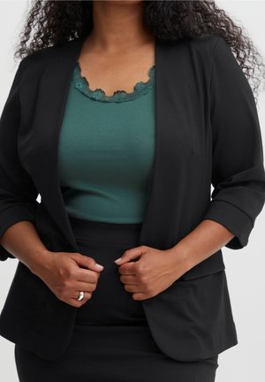 Femme en blazer noir et jupe portant un haut bleu sarcelle orné de dentelle, ajustant son blazer avec les deux mains, bague visible à la main droite.