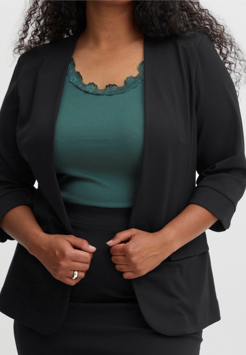 Femme en blazer noir et jupe portant un haut bleu sarcelle orné de dentelle, ajustant son blazer avec les deux mains, bague visible à la main droite.