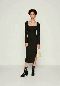 Vestido preto de manga longa ajustado com decote quadrado e fenda lateral, combinado com botas de salto pretas e uma pequena bolsa amarelo-claro.