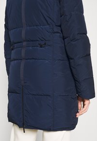 Giubbotto imbottito blu navy con cuciture orizzontali, zip laterale e cordino regolabile in vita per una vestibilità sicura. Texture liscia.